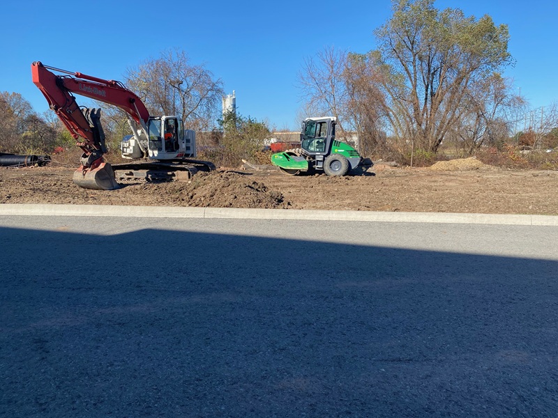 Excavator and compaction equipment grading soil beside a paved roadway
